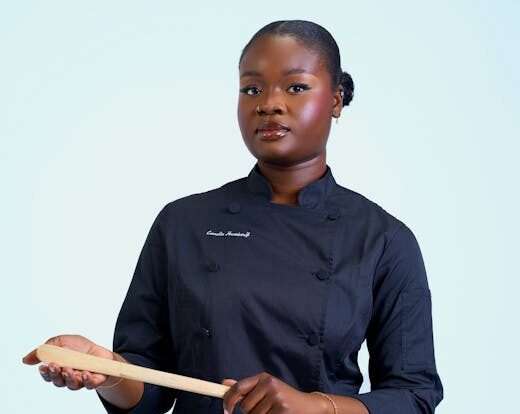 A professional chef wearing a black uniform holding a wooden spoon against a neutral background.